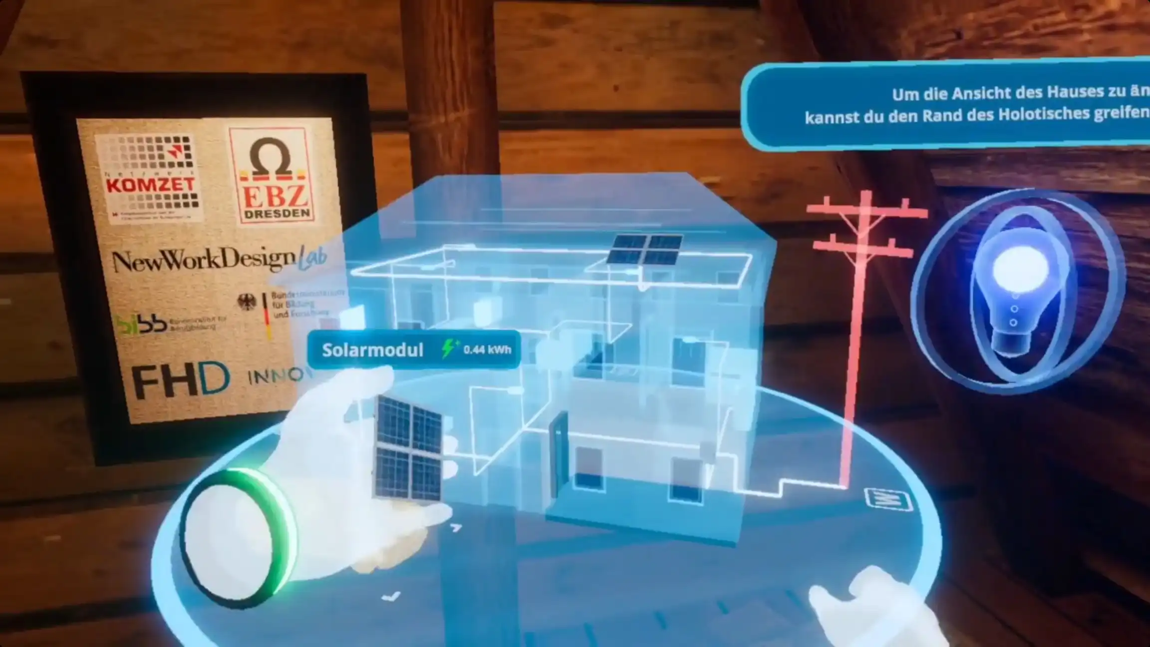 VR-Szene: Eine Hand greift ein Solarmodul mit der Anzeige „0,44 kWh“ und platziert es am holografischen Hausmodell, daneben ein Hinweis zur Bedienung des Holotisches.