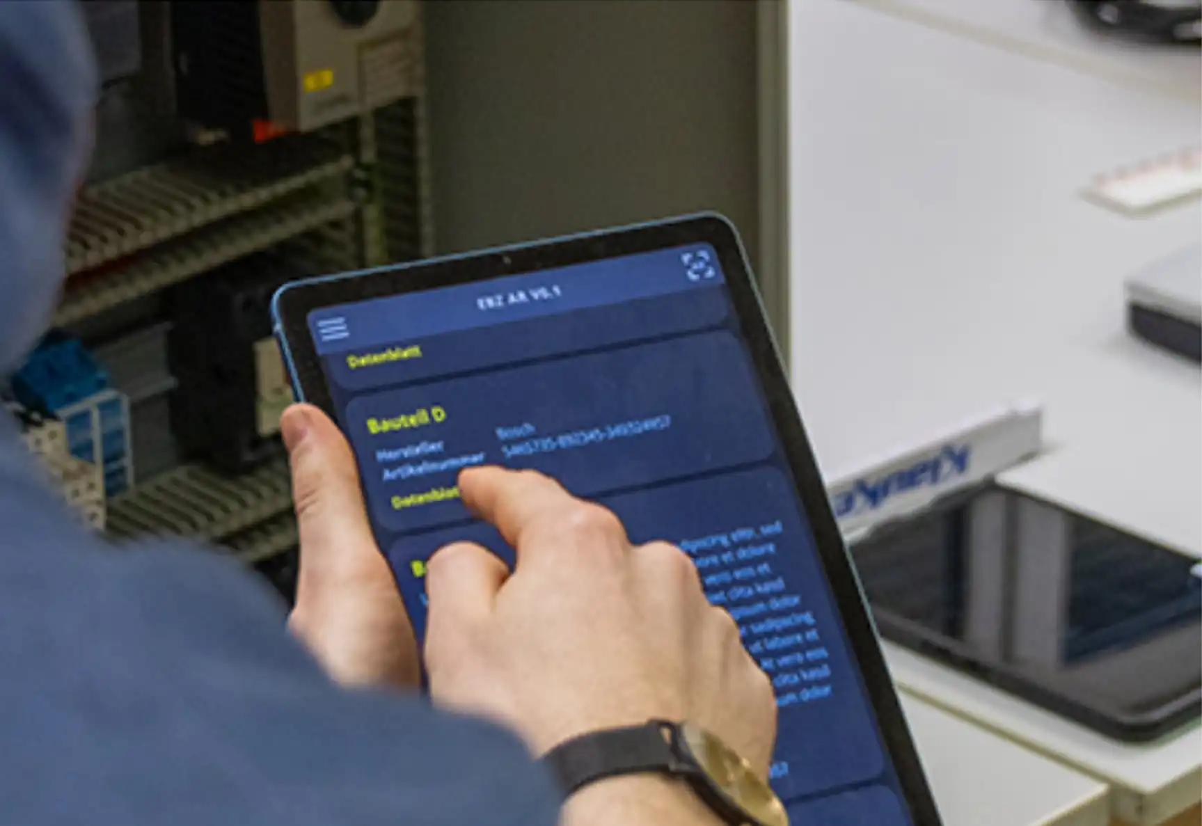 Blick über die Schulter auf ein Tablet mit der App „EBZ AR“: Auf dem Bildschirm wird das Datenblatt eines Bauteils mit Hersteller und Artikelnummer eingeblendet, im Hintergrund ein realer Schaltschrank.