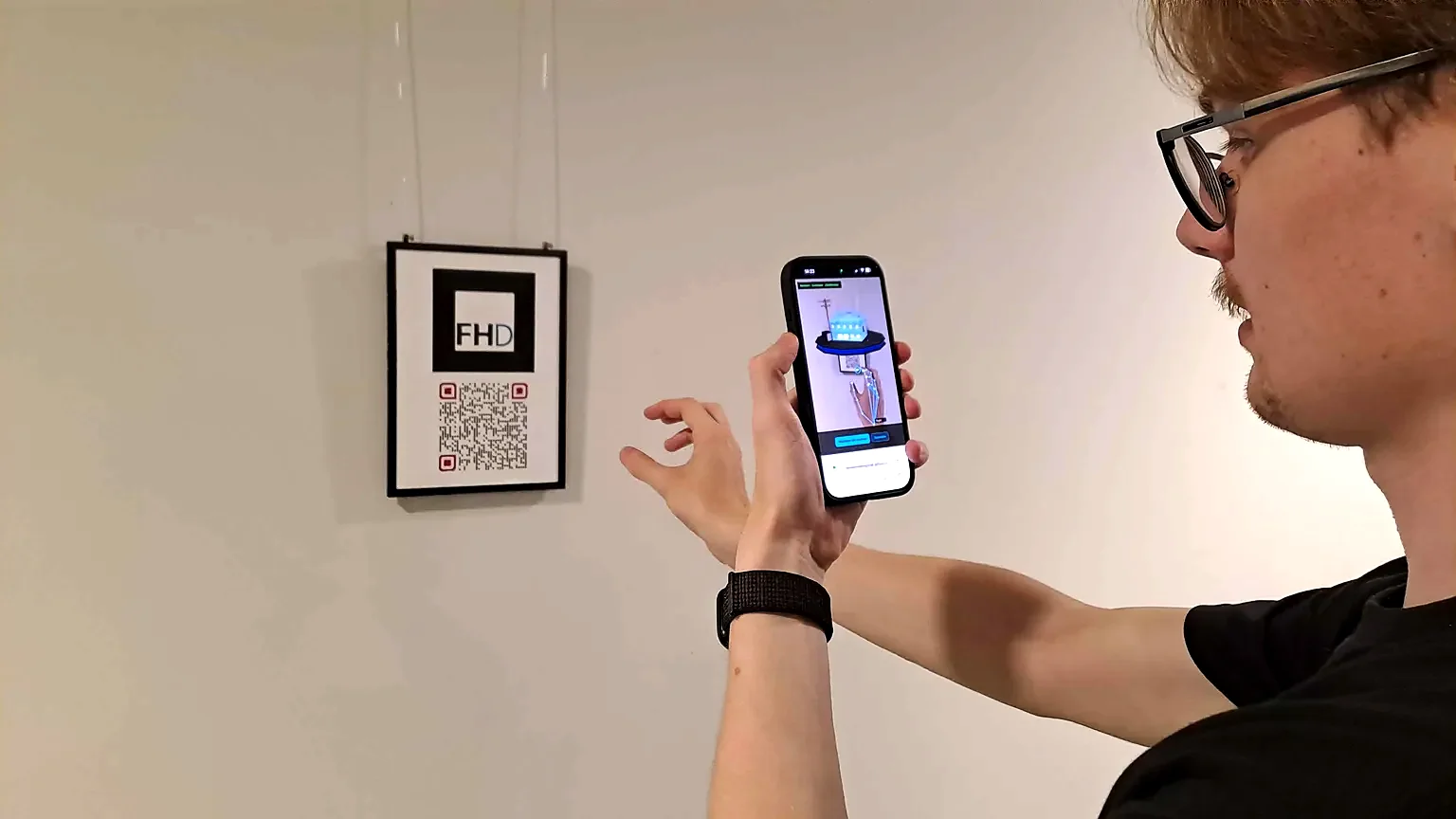 Eine Person hält ein Smartphone vor einen gerahmten QR-Code an einer Wand; auf dem Display erscheint ein blau leuchtendes 3D-Modell über dem Code.
