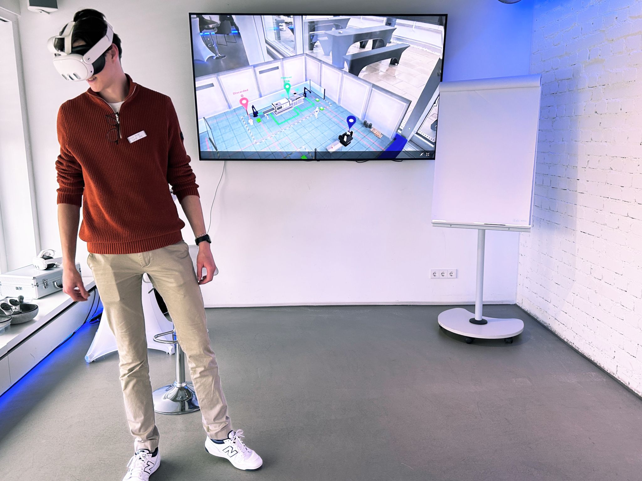 Person mit VR-Brille in einem Showroom; der Monitor an der Wand zeigt die virtuelle ZEISS-Fabrikhalle mit Markierungspunkten.